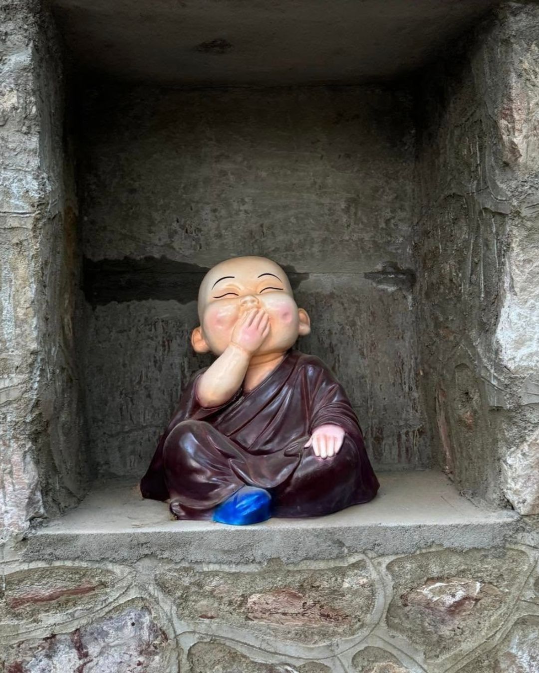 Little monk statue in maroon robe covering mouth speak no evil pose